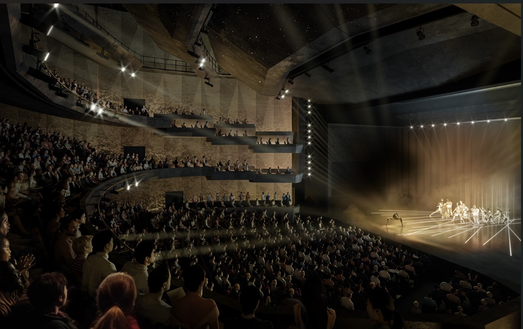 The image is from a high perspective, showing a dark auditorium with up to 2,000 people watching a performance dance performance on stage. There is an indistinguishable number of performers on stage,