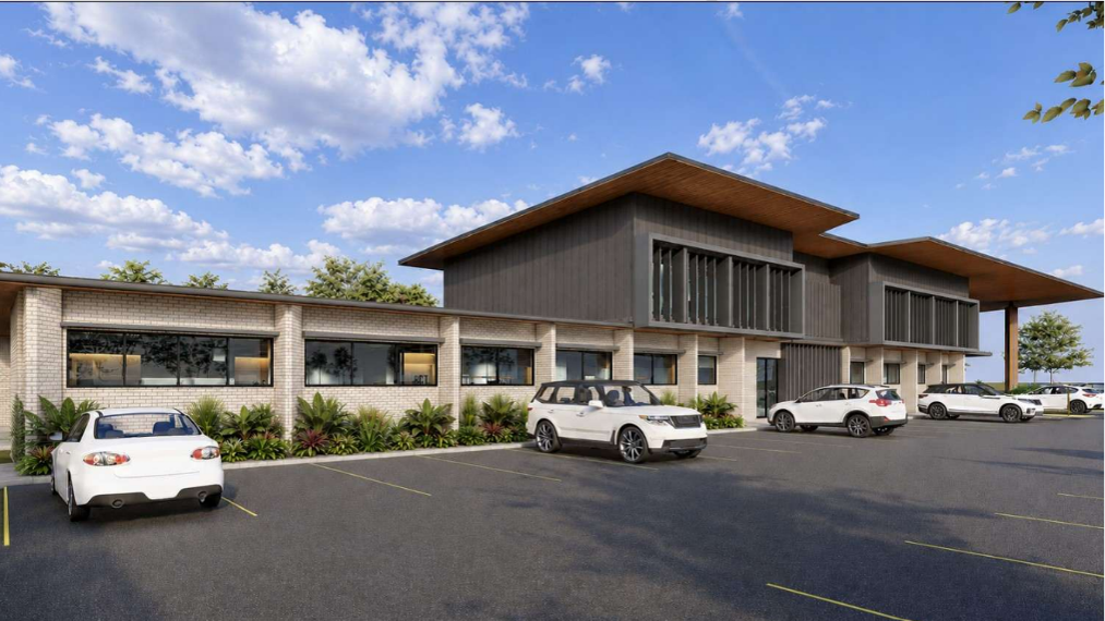 Exterior rendering of a contemporary healthcare building with a mix of light brick and dark cladding, large windows, landscaped garden beds, and a parking area with several parked cars under a blue sky with scattered clouds.