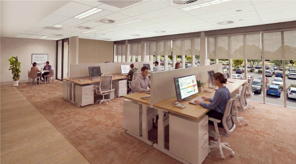 Open-plan office workspace with grouped desks and computer workstations arranged across a carpeted floor. Several people are working at desks separated by low privacy panels. Large windows along one side of the room provide natural light and views of a parking area outside. A small meeting space with a whiteboard and two people is located in the back corner.
