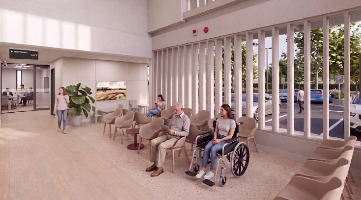 Bright, modern healthcare waiting area with rows of upholstered chairs arranged along large floor-to-ceiling windows overlooking a parking area. Several people are seated, including one person using a wheelchair in the foreground and another reading in a nearby chair. A large potted plant, wall-mounted artwork, and light wood flooring create a welcoming atmosphere. An entrance corridor with directional signage is visible on the left side of the space.
